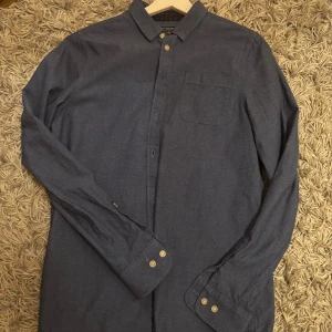 Blå skjorta från Smart With Style - Snygg blå skjorta från Smart With Style med klassisk krage och bröstficka. Skjortan har långa ärmar och knappar framtill samt vid ärmsluten. Materialet känns mjukt och lätt, perfekt för dig som gillar stilrena plagg med enkel design.