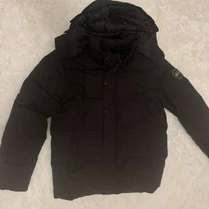 Svart dunjacka från Canada Goose - Svart dunjacka från Canada Goose med stor huva och klassisk logga på ärmen. Jackan har dragkedja och knappar framtill, samt en puffad design som håller dig varm. Perfekt för kalla vinterdagar och har en clean, stilren look.