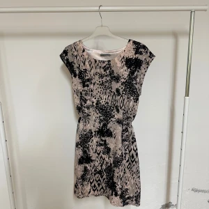 Beige och svart kortklänning Soaked in Luxury - Snygg kortklänning från Soaked in Luxury i beige med svart abstrakt mönster. Klänningen har rund hals, ärmlös design och ett lätt glansigt material som ger en lyxig känsla. Perfekt för dig som gillar att sticka ut med coola prints och enkel passform.