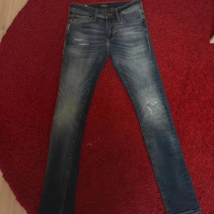 Jack & Jones Skinny Liam jeans blå - Snygga blå skinny jeans från Jack & Jones, modell Liam. Jeansen har slitna detaljer och klassisk femficksdesign. De är tillverkade i stretchig denim för en tight passform och har snygga slitningar på låret och knät.