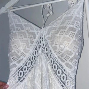 Vit spetsklänning med snörning - Supersöt vit kortklänning med spetsdetaljer och geometriskt mönster. Klänningen har omlott-design framtill, snörning i ryggen och är ärmlös med smala axelband. Perfekt för sommarens alla tillfällen, med snygga volanger och genombruten spets som ger en boho-vibe.