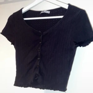 Svart croppad blus från Sister S - Svart croppad blus med korta ärmar och v-ringning från Sister S. Toppen har ribbad struktur, dekorativa knappar framtill och vågiga kanter vid ärmslut och nederkant. Perfekt för dig som gillar en enkel men snygg look.