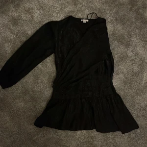 Svart klittrig omlottklänning Gina Tricot - Supersnygg svart klittrig kort omlottklänning från Gina Tricot med långa puffärmar och v-ringning. Klänningen har en lätt glansig yta och markerad midja med volang nertill. Perfekt för dig som gillar en stilren look med lite extra detalj.