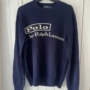 Marinblå stickad tröja Polo Ralph Lauren - Snygg marinblå stickad tröja från Polo Ralph Lauren med stor vit logga framtill. Klassisk rund halsringning och ribbade muddar vid ärmslut och nederkant. Perfekt för dig som gillar stilren och tidlös design.