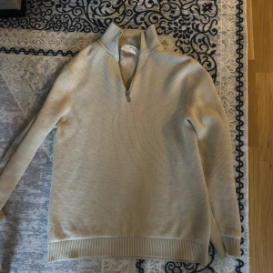 Beige stickad tröja Selected Homme - Beige stickad tröja från Selected Homme med ribbad krage och half zip-dragkedja. Tröjan har lång ärm och breda ribbade muddar vid ärmslut och nederkant. Perfekt för lager-på-lager och chill stil.