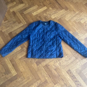 Quiltad blå jacka från Uniqlo XS - Snygg quiltad jacka från Uniqlo i marinblå färg. Jackan har rund hals, lång ärm och dragkedja framtill. Tunn och smidig. Lagom för höst och vår eller som extra lager under annan jacka på vintern. Fint vänt skick. På ena ärmen finns ett vitt litet streck, se bild. 
