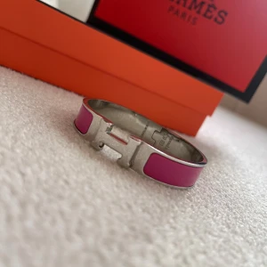 armband i  silver - Säljer ett ikoniskt armband i silverfärgad metall med rosa emaljdetaljer. Armbandet har den klassiska formade låset och en stilren, modern design. Perfekt för dig som vill ha en färgklick på handleden och älskar lyxiga accessoarer.