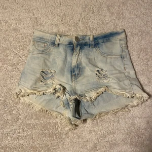 Ljusa jeansshorts - Säljer ett par ljusblå jeansshorts från Gogo Star med hög midja, slitna detaljer och fransig kant. Shortsen har klassisk femficksdesign och knäppning fram. Perfekta för sommaren!
