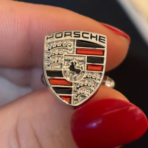 Porsche manschettknapp i silverfärg - Cool manschettknapp med Porsche-logga i silverfärg, dekorerad med svarta och röda detaljer. Sköldformad design med tydlig text och emblem, perfekt för dig som gillar bilar och vill sticka ut med accessoarer.