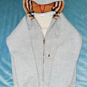 Grå Burberry hoodie med dragkedja - Säljer en grå hoodie från Burberry med klassiskt rutigt foder i huvan. Tröjan har dragkedja framtill, snörning vid huvan och broderad logga på bröstet. Perfekt för dig som vill ha en stilren och lyxig look med en touch av ikoniskt mönster.