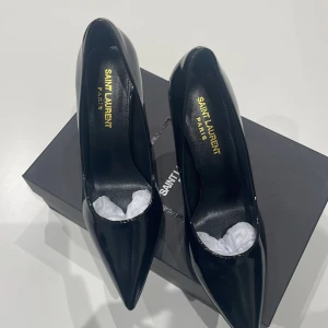 Svarta Saint Laurent pumps med YSL-klack - Säljer ett par svarta pumps från Saint Laurent i blankt skinn med spetsig tå och hög klack i guld med ikoniska YSL-loggan. Snygg och edgy design som verkligen sticker ut. Perfekt för dig som vill ha något extra till outfiten.