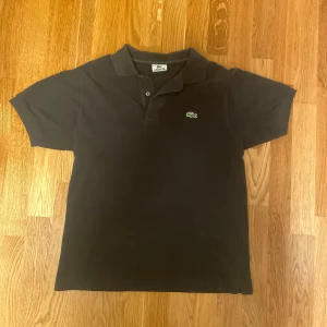 Svart pikétröja från Lacoste - Klassisk svart pikétröja från Lacoste med den ikoniska gröna krokodilloggan på bröstet. Tröjan har korta ärmar, krage och knäppning med två knappar. Perfekt för en clean och sportig stil. Tillverkad i bomull för skön känsla.