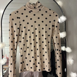 Beige prickig polotröja - Snygg beige polotröja med svarta prickar och lång ärm. Tröjan har en tight passform och är tillverkad i ett mjukt, stretchigt material som sitter skönt mot kroppen. Perfekt att styla med jeans eller kjol för en trendig look.