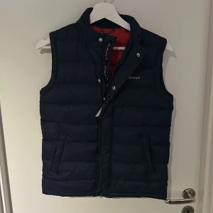 Mörkblå dunväst från GANT - Snygg mörkblå dunväst från GANT med broderad logga på bröstet och röd insida. Västen har ståkrage, dragkedja och tryckknappar framtill samt två sidofickor. Hittade i garderoben och har inte använt på år, därför säljer jag den