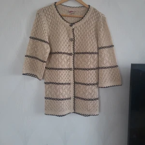 Beige glittrig kofta med svarta detaljer - Snygg beige kofta med glittrigt material och svarta broderade ränder. Koftan har trekvartsärm, rund hals och knappar framtill. Tillverkad av 90% akryl och 10% elastan för en mjuk och stretchig känsla. Perfekt för dig som gillar vintagevibbar och unika detaljer.