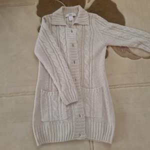 Beige kabelstickad kofta från Ellos - Säljer en beige kabelstickad kofta från Ellos med krage, stora fickor framtill och knappar hela vägen. Koftan är lång och har ribbade muddar vid ärmslut och nederkant. Materialet är 70% akryl och 30% ull, perfekt för kyliga dagar.