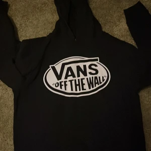 Svart Vans hoodie med stor logga - Svart hoodie från Vans i storlek M ( passar ca 148-156) med stor vit 'Vans Off The Wall'-logga på bröstet. Väldigt bra kvalite. Klassisk huva och långa ärmar, perfekt för dig som gillar streetwear och vill ha en enkel men snygg look. Materialet känns mjukt och bekvämt. 
