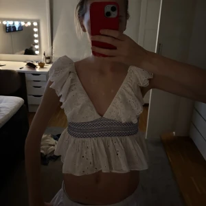 Vit volangig blus med broderi - Supersöt vit blus med broderade hål och volangiga axlar. Djup v-ringning fram och bak, samt smockad midja med blått broderi. Luftig och perfekt för varma dagar, med romantisk känsla och ärmlös design.