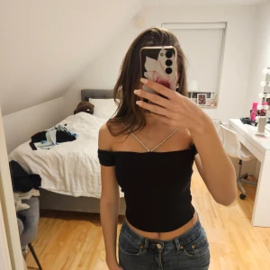 Svart croppad offshoulder topp - Säljer en svart croppad offshoulder topp med smala glittriga axelband som korsas framtill. Toppen är tight och har en snygg, enkel design som passar perfekt till jeans eller kjol. Materialet känns stretchigt och bekvämt.