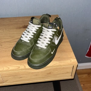 Nike Air Force 1 gröna sneakers - Säljer ett par feta Nike Air Force 1 sneakers i olivgrönt skinn med vita swoosh-loggor och vita snören. Skorna har klassisk rund tå, platt sula och snygga detaljer med svart och gult på hälen. Perfekta för dig som vill sticka ut med stilrena kicks.