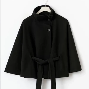 Svart Gina Tricot Belt Cape Jacket - Snygg svart poncho liknande kappa från Gina Tricot. Den har breda ärmar, hög krage och knappar framtill. Ponchon har ett matchande bälte i midjan som ger en clean och modern siluett. Perfekt för dig som gillar minimalistisk stil och vill ha något som sticker ut med sin coola form. Ångrade mitt köp men hade inte tagit med kvitto, prislapp finns kvar
