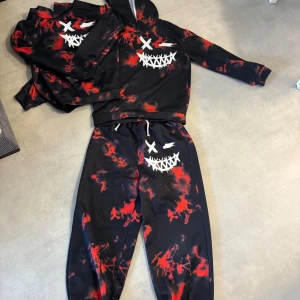 Svart och röd hoodie från Shein - Cool hoodie från Shein med svart och röd batikmönster och ett vitt smiley-ansikte tryckt på bröstet. Tröjan har huva, långa ärmar och är gjord i mjukt syntetmaterial. Byxorna och ena tröjan är storlek 11 år medan den andra är storlek 10 år. Kolla gärna in mina andra artiklar!