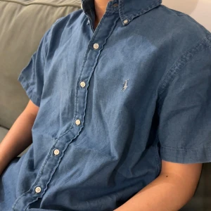 Blå kortärmad skjorta från Ralph Lauren - Snygg blå skjorta från Ralph Lauren i modellen Indigo Oxford. Skjortan har korta ärmar, klassisk button-down krage och vita knappar framtill. Materialet är mjuk bomull och den har den ikoniska broderade loggan på bröstet. Perfekt för en chill och stilren look.