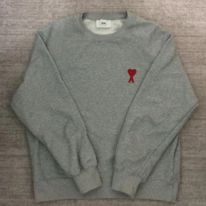 Grå sweatshirt från Ami Paris - Grå sweatshirt från Ami Paris med röd broderad hjärtlogga och bokstaven A på bröstet. Klassisk rund halsringning, ribbade muddar och mjuk bomullskänsla. Stort Ami-tryck på framsidan. Perfekt för en clean och trendig look.