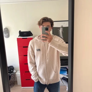 Beige hoodie från BVALDI med dragkedja - Mysig beige hoodie från BVALDI i fluffigt stickat material. Tröjan har huva med snörning, hel dragkedja och broderad BV-logga på bröstet. Perfekt för dig som gillar en chill och avslappnad stil.