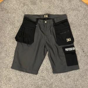 Arbetsshorts XL Bygg grå/svarta M - Grymma arbetsshorts från XL Bygg i grått och svart med flera stora fickor, dragkedja och coola detaljer. Perfekta för dig som vill ha praktiska och slitstarka shorts med plats för verktyg. Märke på fickan och midja med knappstängning. Snygga arbetsshorts som är perfekt för dig som gillar att hänga i verkstan och snickra. Jättefint skick på dem och har ej använts så hilma mycket. Hör av er om ni har frågor!