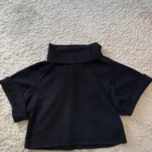 Svart croppad polotröja - Svart polotröja med croppad passform och korta ärmar. Tröjan har en bred, uppvikt krage och är tillverkad i ett mjukt stickat material som känns skönt mot huden. Perfekt för dig som gillar en clean och stilren look.