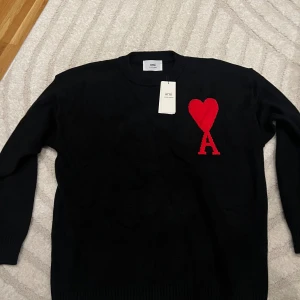 Svart stickad tröja från Ami - Svart stickad tröja från Ami med rund hals och lång ärm. På bröstet finns ett stort rött hjärta och bokstaven A som broderad detalj. Tröjan har ribbade muddar vid ärmslut och nederkant, vilket ger en snygg och clean look.