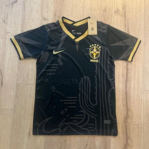 Svart Brasil Nike fotbollströja S - Snygg svart Brasilien fotbollströja från Nike med guldiga detaljer och emblem. Tröjan har V-ringad krage med guldkant, korta ärmar med guldrand och subtilt grafiskt mönster på framsidan. Tillverkad i lätt, ventilerande Dri-FIT material som passar perfekt för träning eller match.