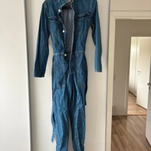Blå denim jumpsuit med bälte - Säljer en blå denim jumpsuit med långa ärmar och flera stora fickor både fram och bak. Jumpsuiten har knappar framtill och ett matchande bälte i midjan för att justera passformen. Perfekt för dig som gillar en cool och avslappnad stil.