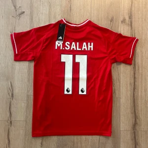 Liverpool Salah tröja Adidas S - Riktigt snygg Liverpool fotbollströja från Adidas i röd färg med vita detaljer. Tryck med M. Salah och nummer 11 på ryggen, Premier League-logga, samt klubbmärke och sponsor framtill. Slim fit, kortärmad och tillverkad i lätt polyester med Aeroready-teknologi.
