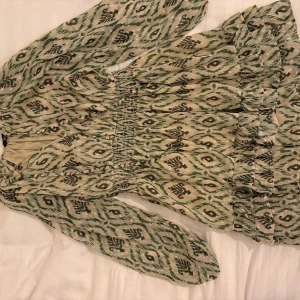 Grön klänning zara - Säljer en kortklänning med grönt och beige mönster, smockad midja och volangdetaljer nedtill. Klänningen har långa ärmar och ett unikt, bohemiskt mönster.