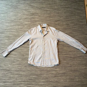 Randig slim fit skjorta från H&M - Snygg randig skjorta från H&M i slim fit-modell. Skjortan är ljusblå med vita tunna ränder, har klassisk krage och långa ärmar med knappar vid manschetterna. Perfekt för dig som gillar stilrena och fräscha plagg. Materialet är mjuk bomull.