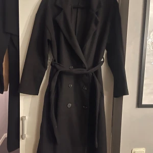 Svart kappa med bälte och knappar - Säljer en svart kappa med klassisk krage, knappar framtill och ett matchande bälte i midjan. Långärmad och med stilren design, perfekt för dig som gillar en clean look. Passar dig som vill ha en tidlös jacka med snygg siluett.