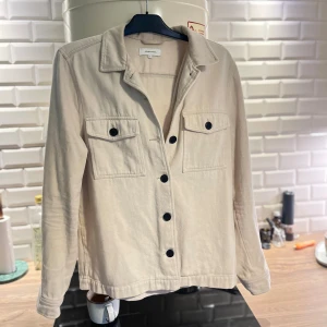 Beige overshirt från Studiototal - Snygg beige overshirt från Stradivarius med klassisk krage och svarta knappar framtill. Två bröstfickor med lock och knapp ger en cool touch. Tillverkad i mjuk bomull, perfekt för lager-på-lager. Långärmad och med avslappnad passform.