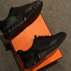 Svarta sneakers med chunky sula - Säljer ett par helsvarta sneakers med chunky, räfflad sula och coola detaljer på sidan. Ovandelen är i mesh och syntetmaterial, vilket ger en modern och sportig vibe. Skorna har snörning och en rund tå, perfekta för dig som gillar stilrena och edgy sneakers.