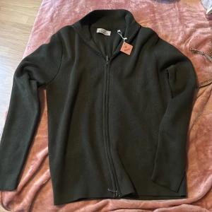 Mörkgrön kofta från Glenn Strömberg - Snygg mörkgrön kofta från Glenn Strömberg i storlek M. Tröjan har hel dragkedja framtill, ribbad krage och långa ärmar. Materialet är stickat och känns mjukt och bekvämt. Perfekt för lager-på-lager under kyliga dagar.