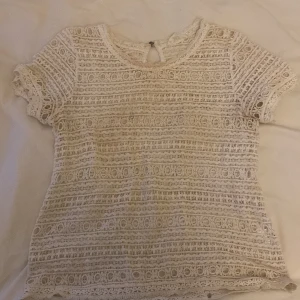 Vit spetsblus med kort ärm - Säljer en söt vit blus med korta ärmar i spets och broderade detaljer. Blusen har rund halsringning och ett litet knappslå i nacken. Perfekt för dig som gillar romantisk och feminin stil med mycket detaljer.