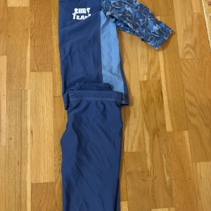 Blå badbyxor Surf Team - Snygga blå badbyxor med elastisk midja och enkel design. Perfekta för stranden eller poolen, med lätt och snabbtorkande material. Matchar med Surf Team-tröja för en cool look. Storlek 110/116 från Kappahl 