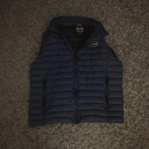 Mörkblå dunväst från Colmar - Snygg mörkblå dunväst från Colmar Originals med quiltad design och dragkedja framtill. Västen har en rundad krage och två sidofickor med dragkedja. Perfekt för lager-på-lager och har en stilren look med Colmar-logga på bröstet. Storlek S passar även M.