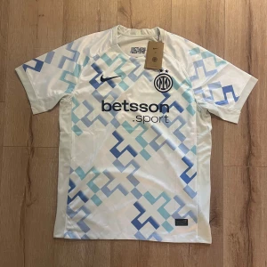 Inter Milan vit och blå fotbollströja Nike - Snygg Inter Milan fotbollströja från Nike i vit med geometriskt mönster i olika blå toner. Tryckt klubbmärke och sponsor på bröstet, samt Nike-logga. Kortärmad modell i lätt och ventilerande polyester, perfekt för match eller träning.