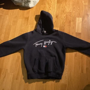 Mörkblå hoodie från Tommy Hilfiger - Säljer en mörkblå hoodie från Tommy Hilfiger med vit broderad logga och klassisk flagga på bröstet. Tröjan har huva med metallöljetter, känguruficka och långa ärmar. Perfekt för dig som gillar stilren streetwear.