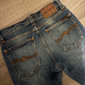 Blå raka jeans från Nudie Jeans - Säljer ett par blå raka jeans från Nudie Jeans med klassisk orange söm och vågig brodering på bakfickorna. Jeansen har slitningar och tvättade detaljer, samt rå kant vid benslut. Perfekt passform för dig som gillar raka ben och snygg streetstil.