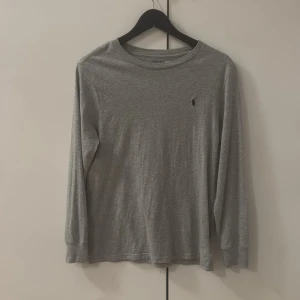 Grå longsleeve tröja Polo Ralph Lauren - En stilren grå långärmad tröja från Polo Ralph Lauren med klassisk rund halsringning och den ikoniska lilla broderade loggan på bröstet. Tillverkad i mjuk bomull som är skön mot huden. Perfekt för lager-på-lager och enkel att matcha.
