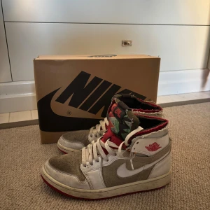 Nike Air Jordan 1 Zoom Air CMFT 41 - Nike Air Jordan 1 Zoom Air CMFT sneakers i vitt, grått och rött med högt skaft. Skorna har snörning, röd innersida och detaljerad tunga med mönster. Swoosh-logga i vitt på sidan och röd yttersula. Materialmix av skinn och mocka för en cool streetstil.