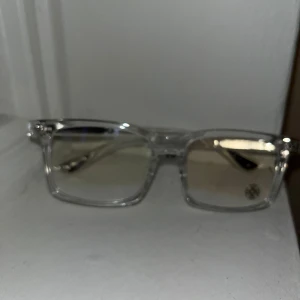 Transparenta glasögon från chrome hearts anti blue - Snygga glasögon med transparent rektangulär båge från chrome hearts. Glasen har en lätt toning och skalmarna är dekorerade med små silverdetaljer. Perfekt för dig som gillar stilrena och moderna accessoarer.